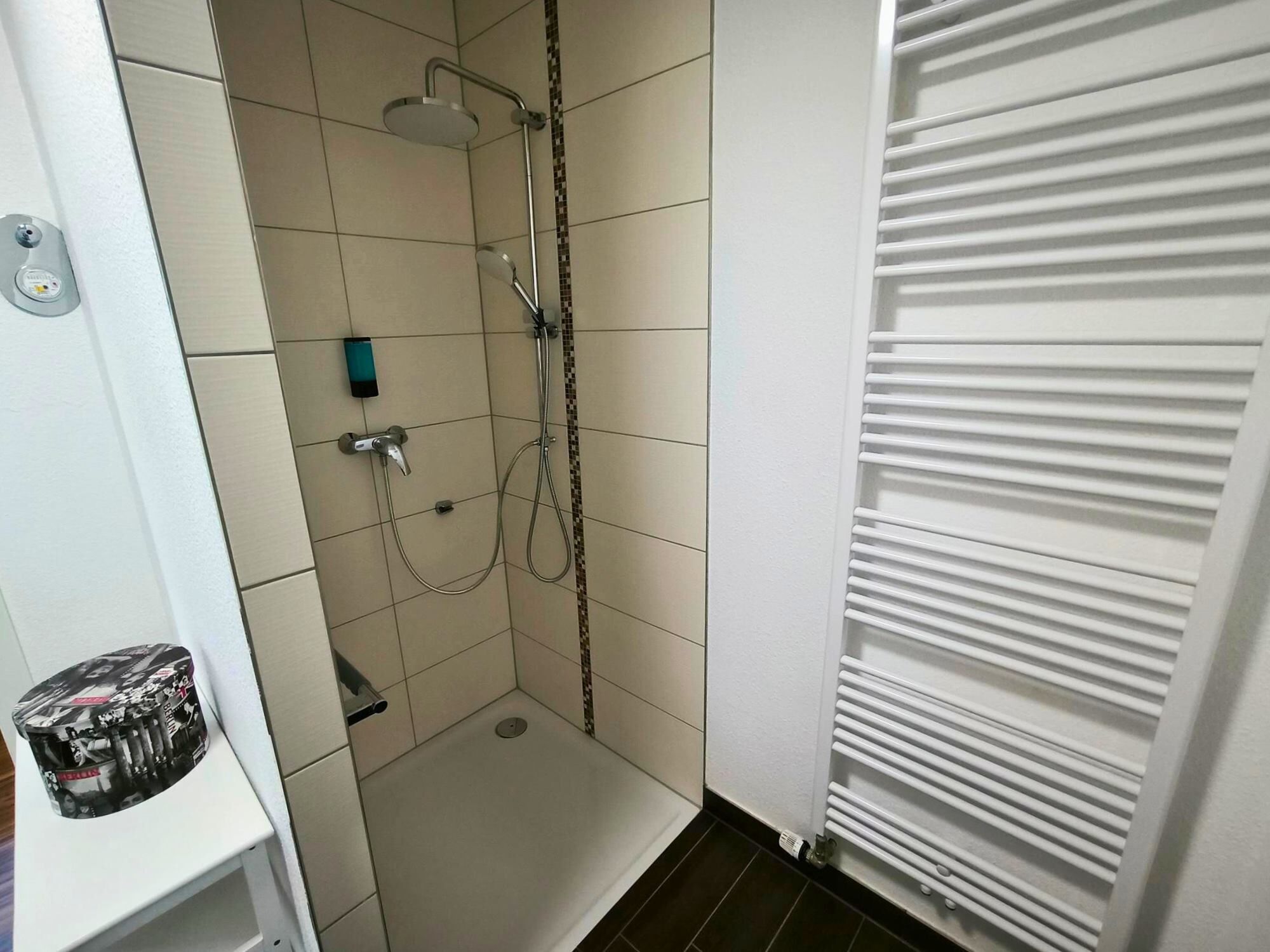 A shower with a handheld showerhead and a wall-mounted faucet. There is a small shelf with a decorative item and a bottle.