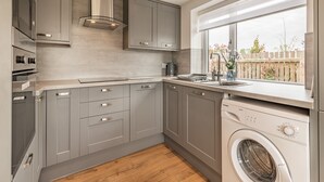 A modern kitchen with grey cabinets, a stainless steel range hood, a built-in dishwasher, and a window with a view of a wooden fence. - 3 bedroom accommodation in Church Broughton, near Derby (Church Broughton, near Derby)