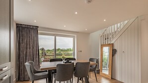 A modern dining area with a wooden table and chairs, large windows with curtains, and a view of greenery outside. - 3 bedroom accommodation in Church Broughton, near Derby (Church Broughton, near Derby)
