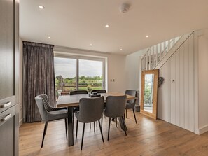 A modern dining area with a wooden table and chairs, large windows with curtains, and a view of greenery outside. - 3 bedroom accommodation in Church Broughton, near Derby (Church Broughton, near Derby)