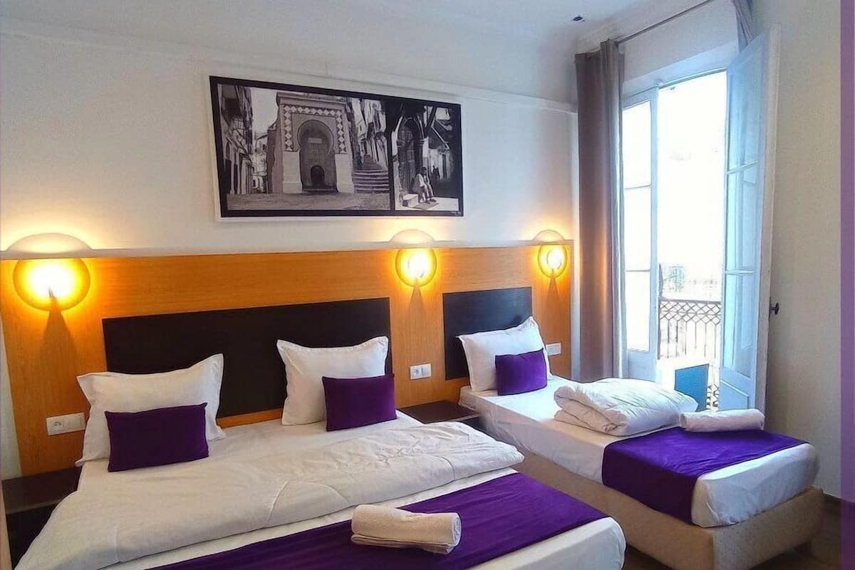 A hotel room with two beds, each with a headboard, white bedding, and purple accents. There is a framed picture above the beds and a window with curtains.