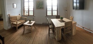 Una habitación con piso de madera, un sofá de madera, una pequeña mesa con una planta, una mesa de comedor con sillas y un televisor montado en la pared.