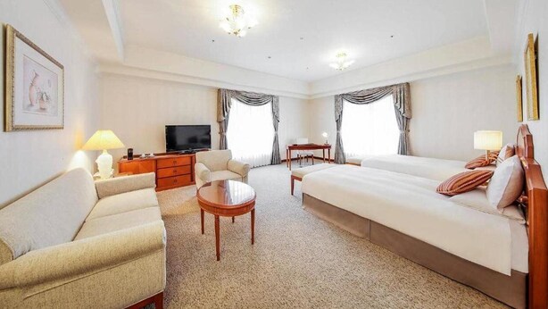 Una habitación de hotel con cama, sofá, escritorio, televisión y dos ventanas con cortinas.