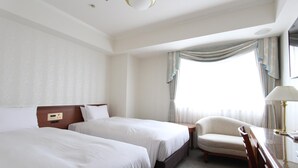 Habitación de hotel con dos camas individuales, una mesita de noche de madera, una silla y un escritorio con lámpara.