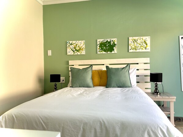 Una cama bien hecha con ropa blanca y almohadas verdes y amarillas, cabecera blanca y dos mesitas de noche con lámparas negras.