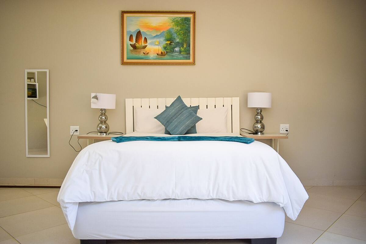 A hotel room with a large bed, a painting of a landscape above it, a mirror on the left, and two bedside lamps.