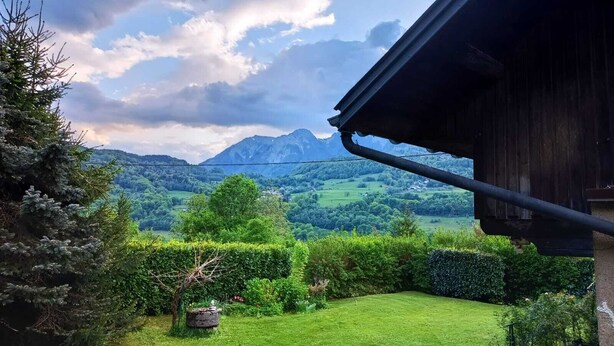 A view from a wooden building with a metal roof, overlooking a lush green garden and a mountainous landscape. - Your Nomadic Room: Africa (MARTHOD)