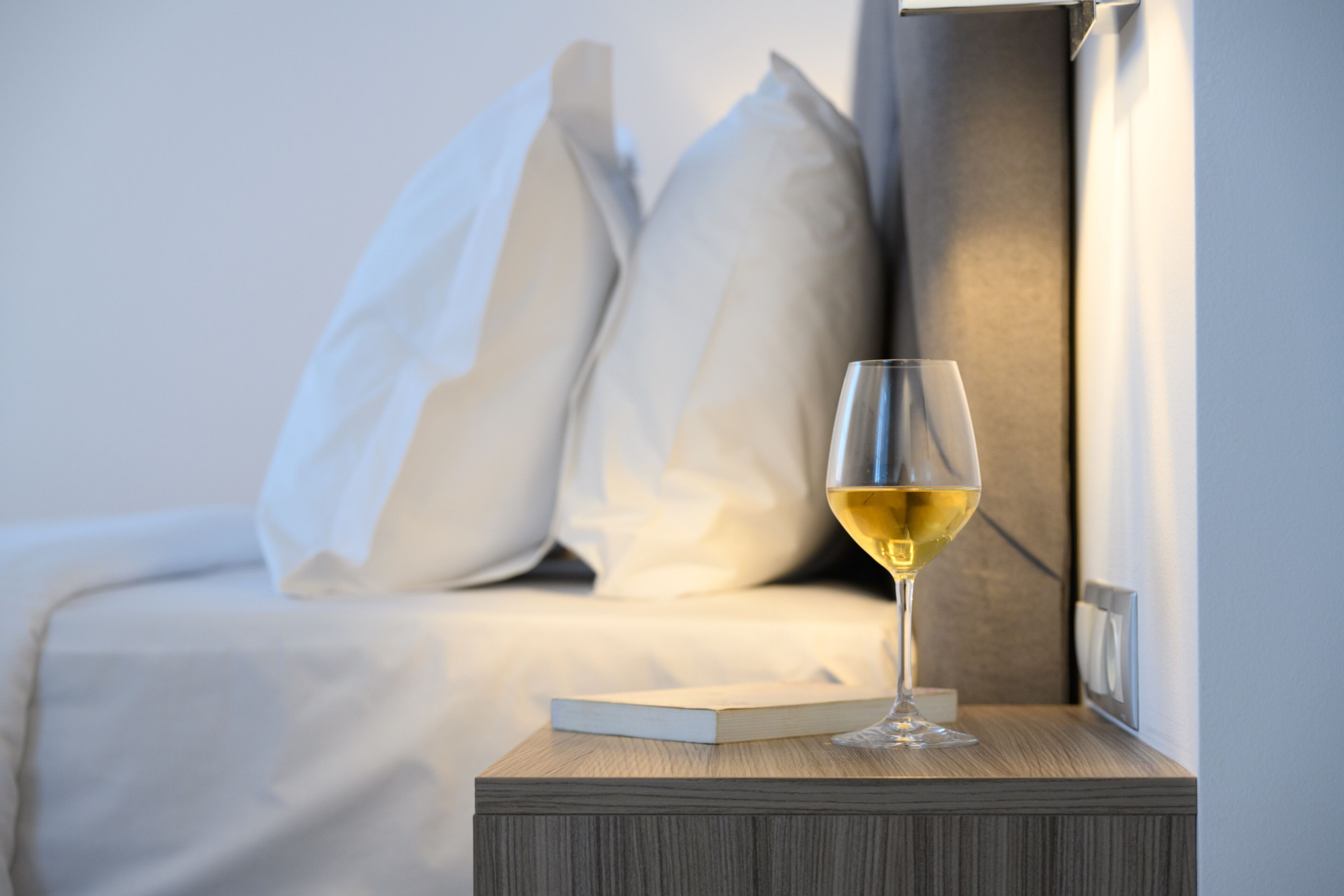 Un lit bien fait, avec du linge de lit blanc, une table de chevet sur laquelle se trouve un verre de vin blanc et un livre.