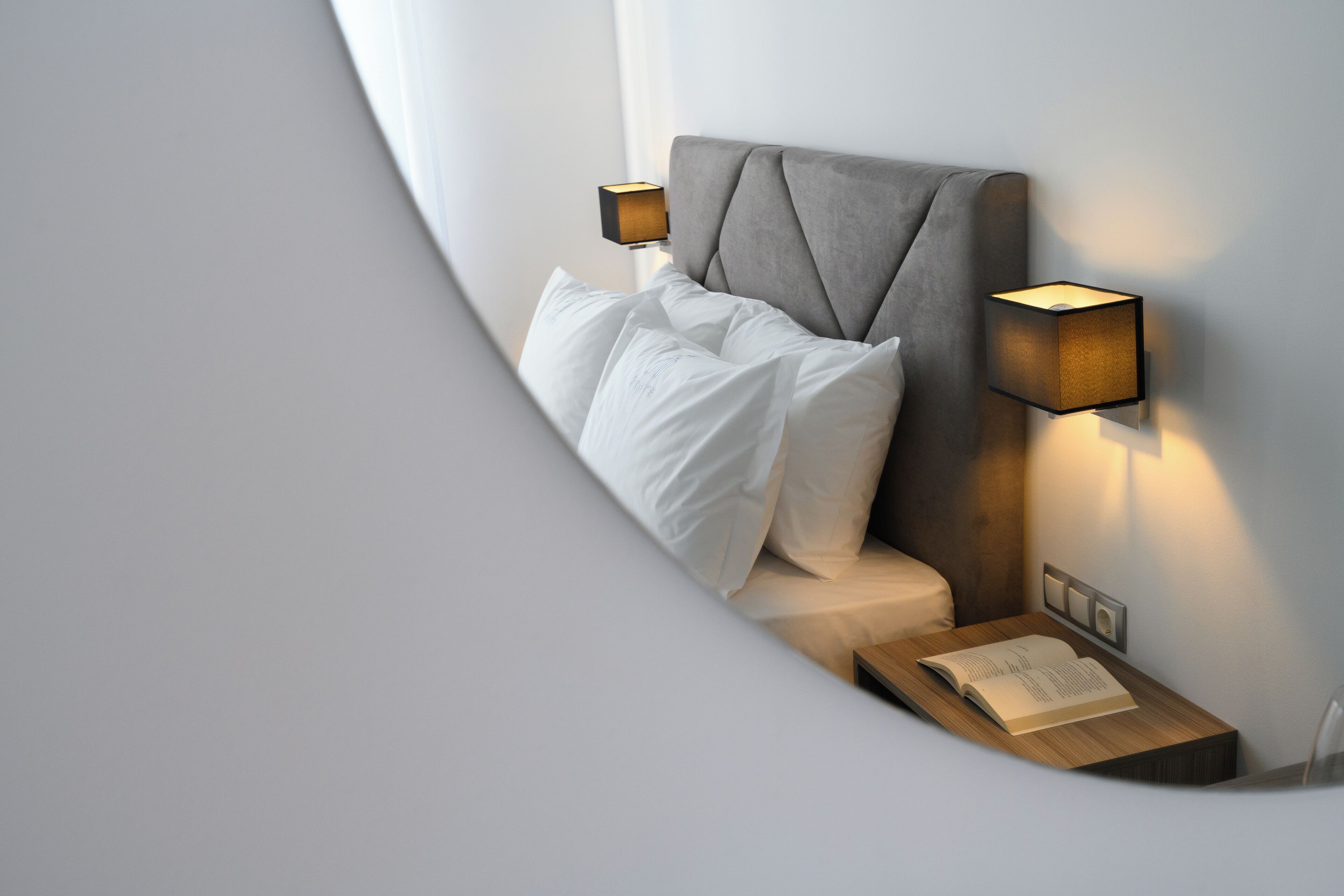 Un dormitorio moderno con una cama bien tendida, dos lámparas de noche fijadas a la pared y una mesita de noche con un libro abierto.