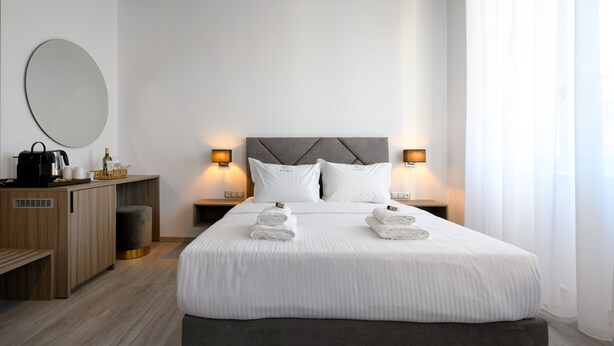 Una habitación de hotel moderna con una cama grande, dos mesitas de noche con lámparas, un escritorio de madera, un espejo redondo y una pequeña zona de estar.