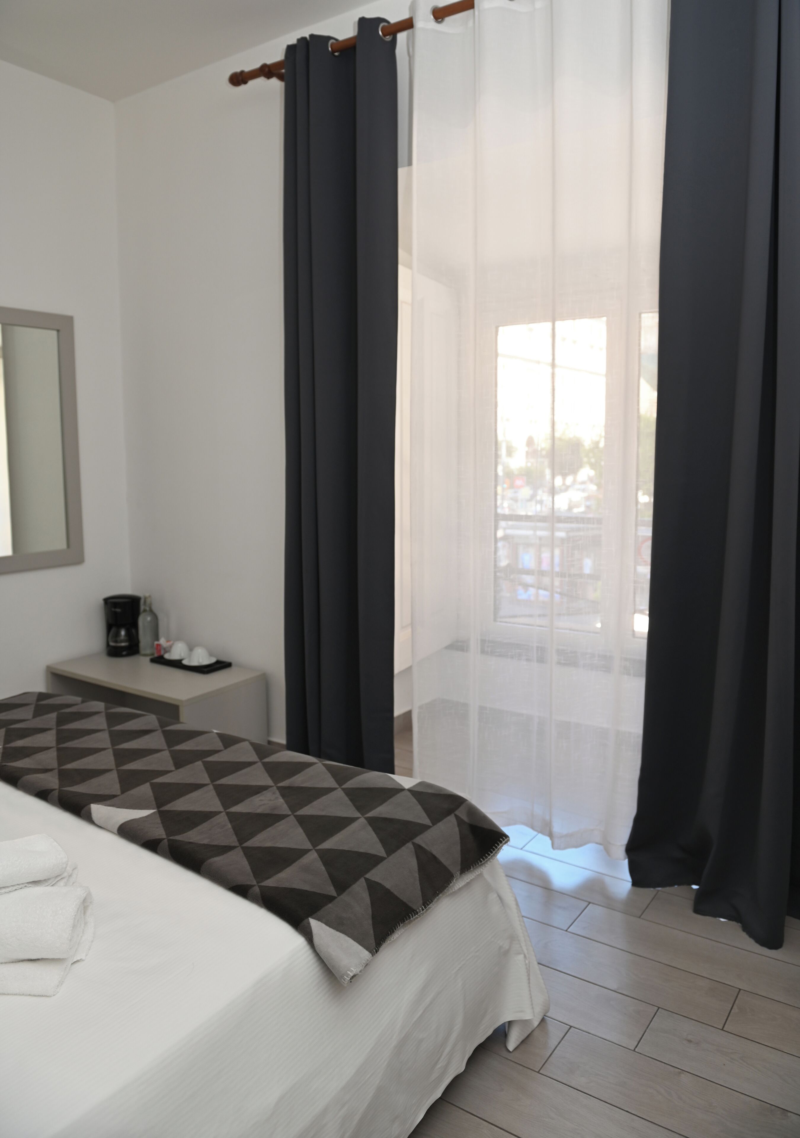 A hotel room with a bed, a bedside table with a coffee maker, and a window with sheer curtains.