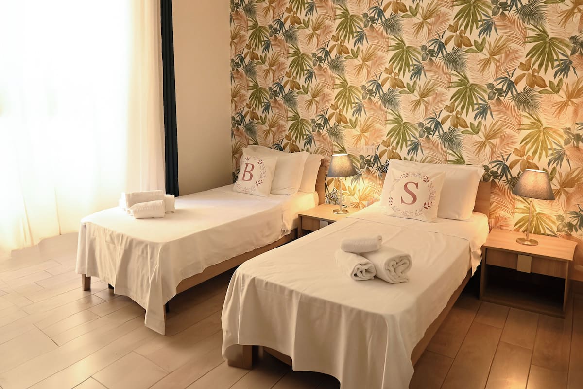 A hotel room with two single beds, each with a white bedspread and matching pillows. The room features a tropical leaf-patterned wallpaper and a wooden floor.