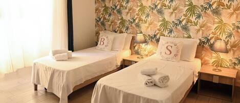 A hotel room with two single beds, each with a white bedspread and matching pillows. The room features a tropical leaf-patterned wallpaper and a wooden floor.