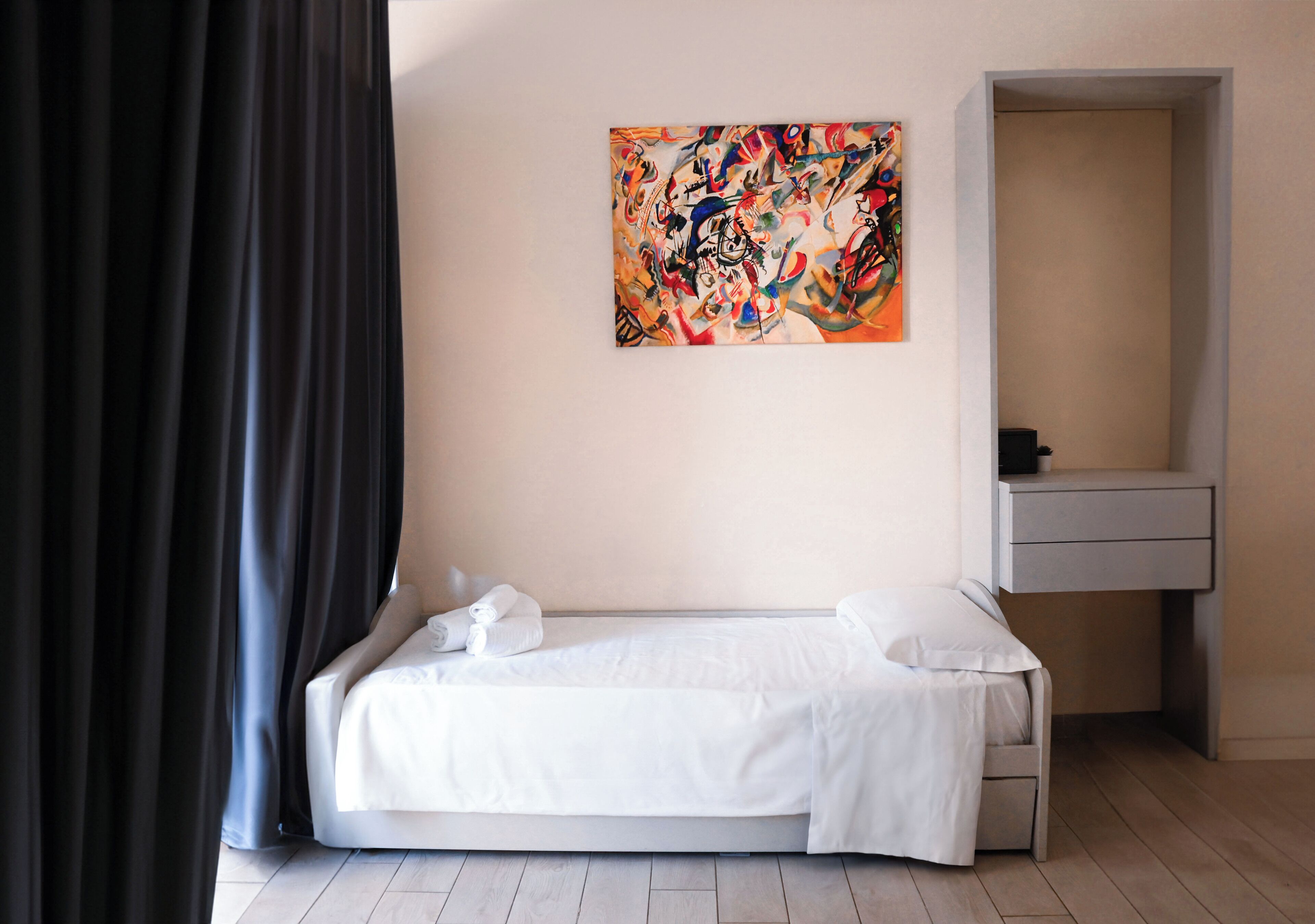 A minimalist hotel room with a single bed, a small bedside table, and a wall-mounted abstract painting.