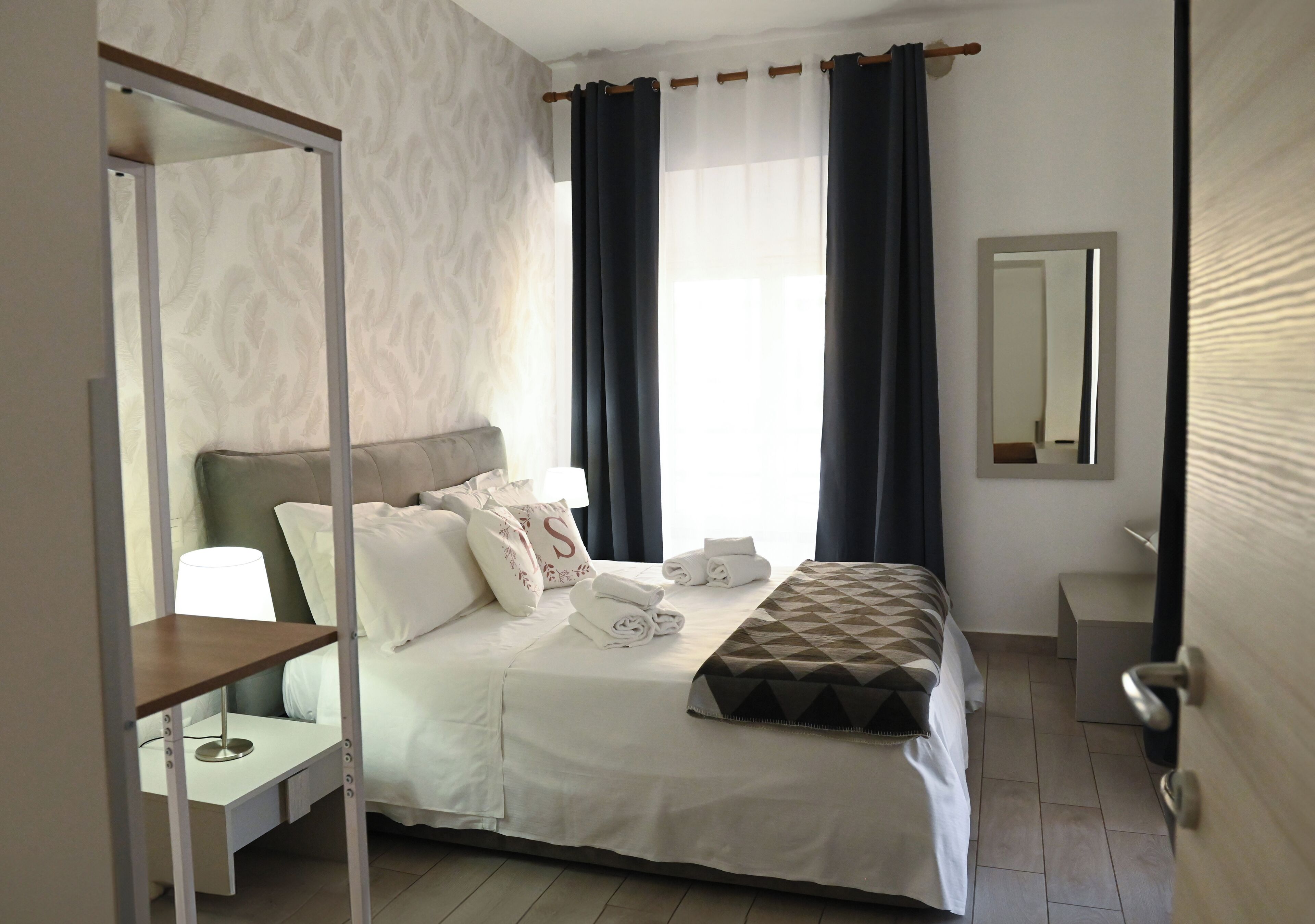 A hotel room with a bed, a bedside table with a lamp, a mirror, and a window with curtains.