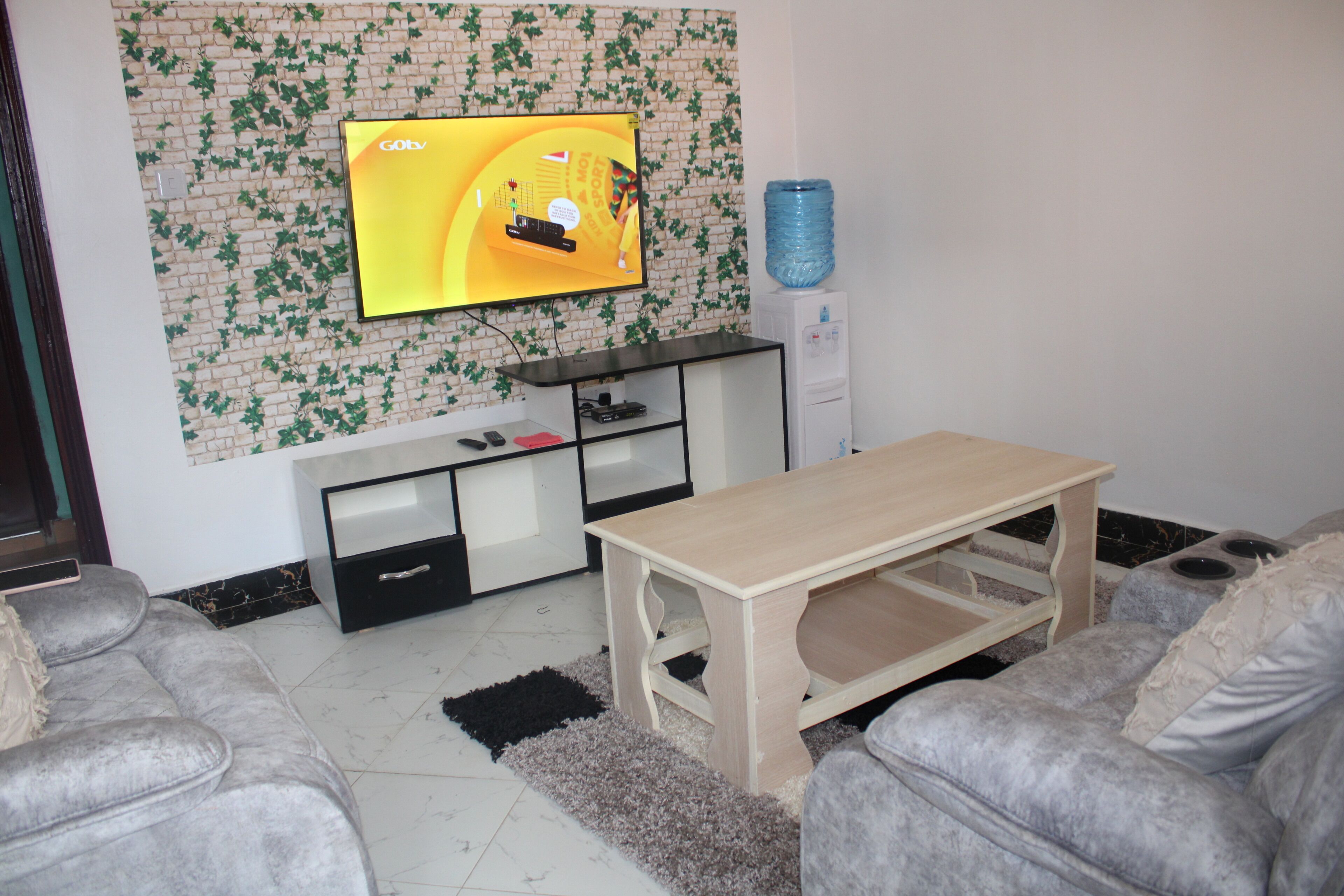 A living room with a flat-screen TV mounted on a brick-patterned wall, a wooden coffee table, grey sofas, and a water dispenser.