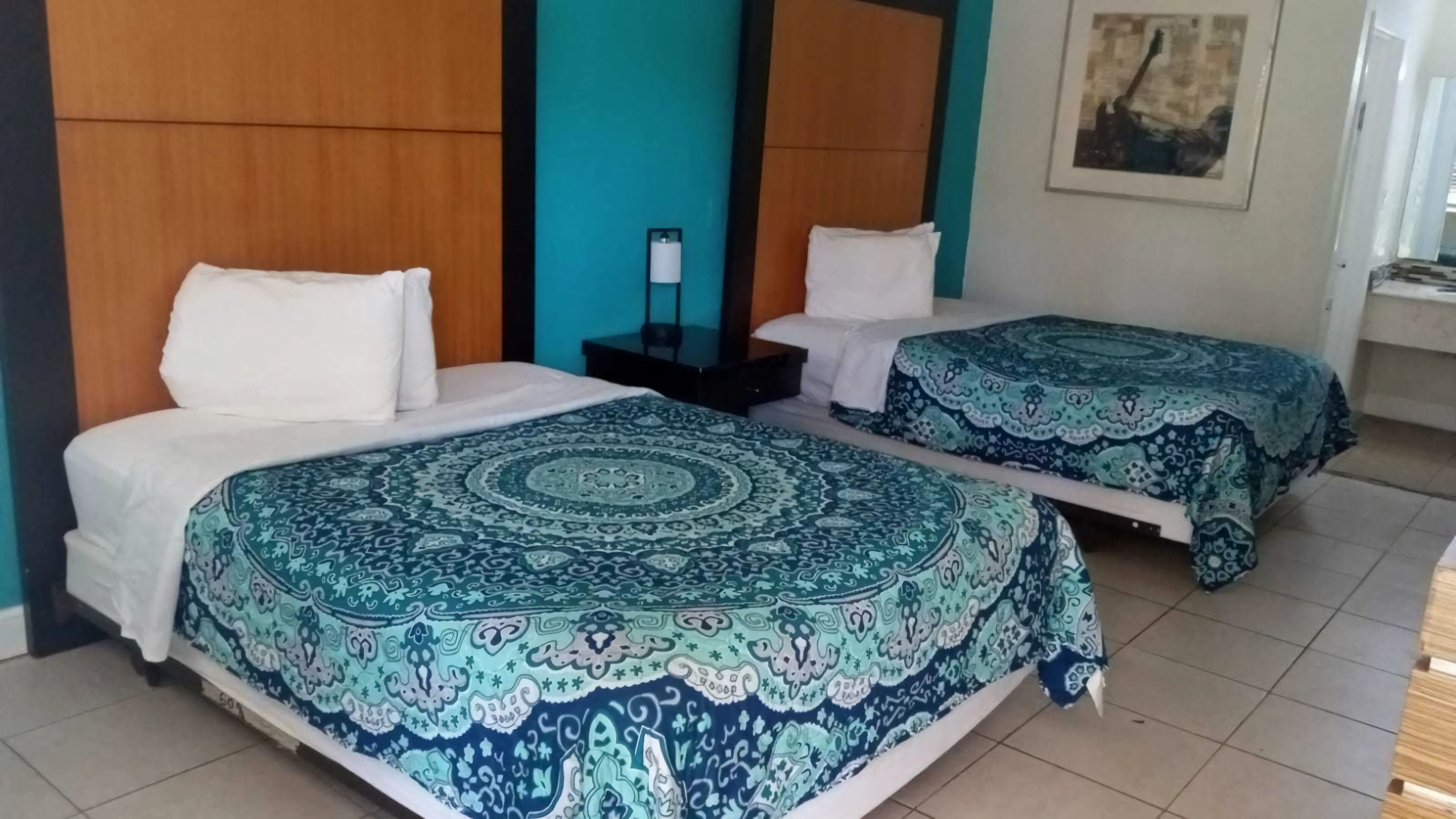 Habitación de hotel con dos camas, cada una con una colcha estampada y almohadas blancas. Hay una mesita entre las camas con una lámpara encima.