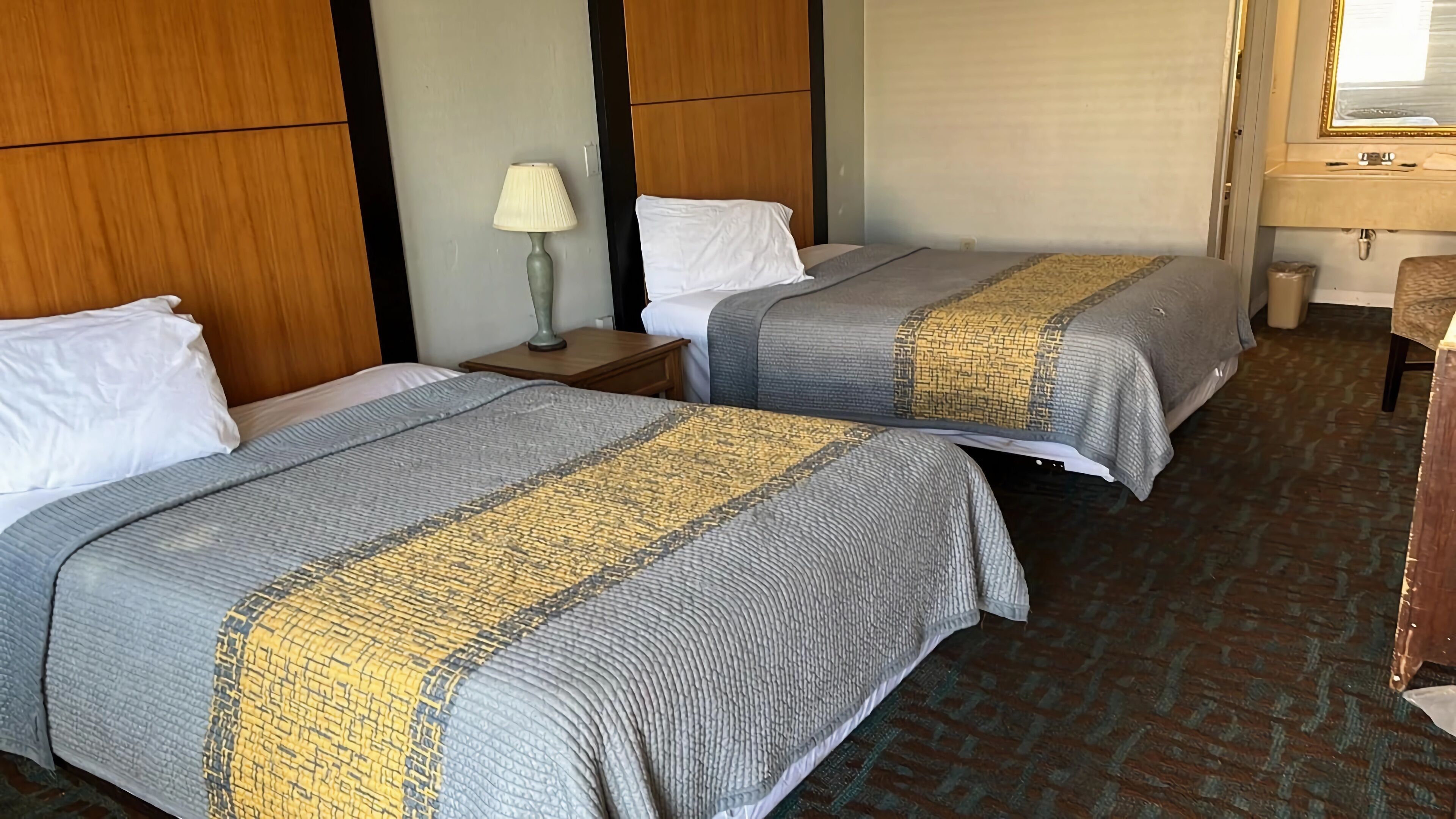 Two beds with grey and yellow striped bedding, a wooden headboard, a nightstand with a lamp, and a bathroom with a sink and mirror.