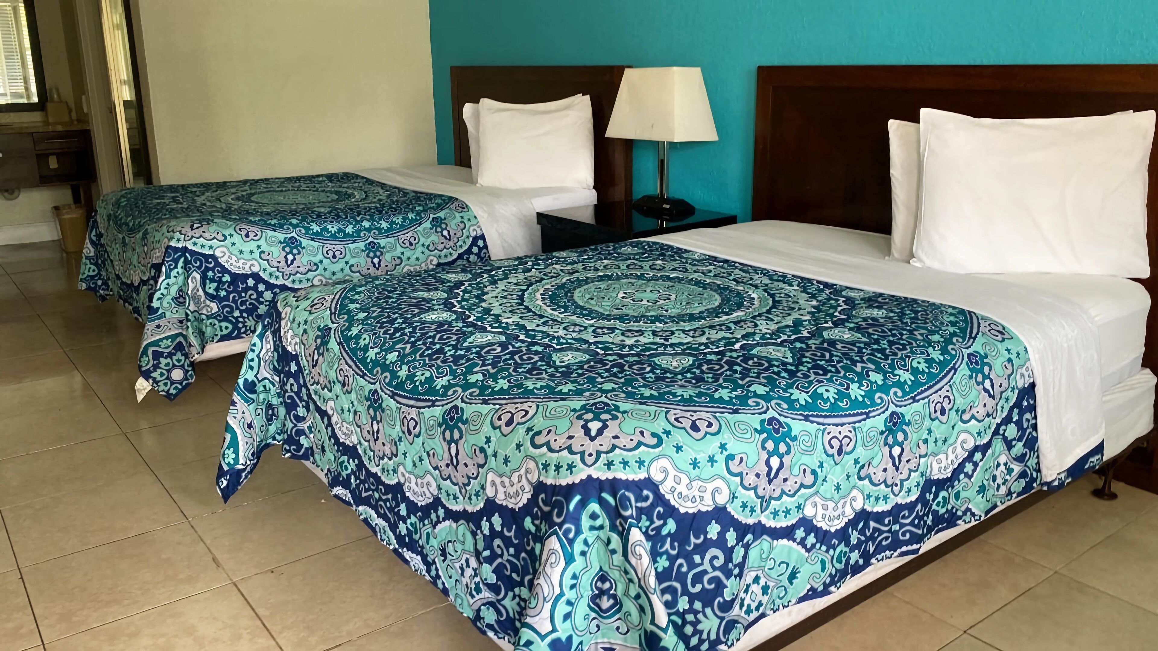 A hotel room with two beds, each with a patterned bedspread, a nightstand with a lamp, and a window with blinds.