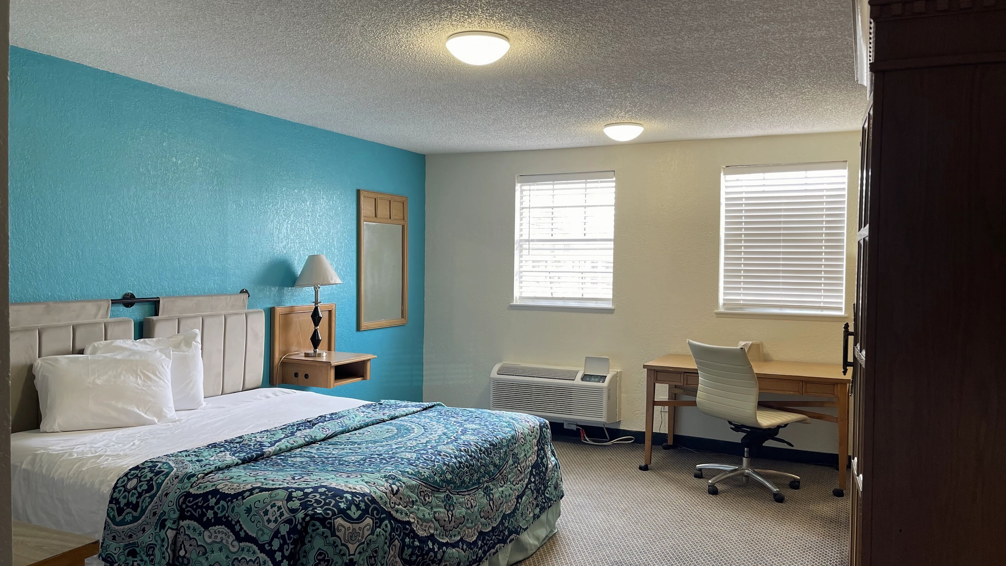 Una habitación de hotel con cama, escritorio, silla, lámpara y dos ventanas.