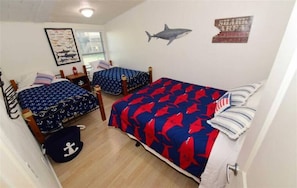 A bedroom with two beds, one with a shark-themed bedspread and the other with an anchor pattern. There is a framed picture on the wall and a shark decal above the beds.