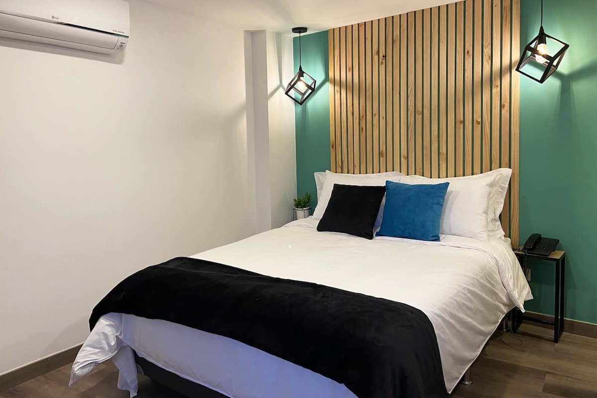 A modern bedroom with a wooden slat headboard, a white bed with black and blue pillows, a black bedside table with a phone, and an air conditioning unit on the wall.