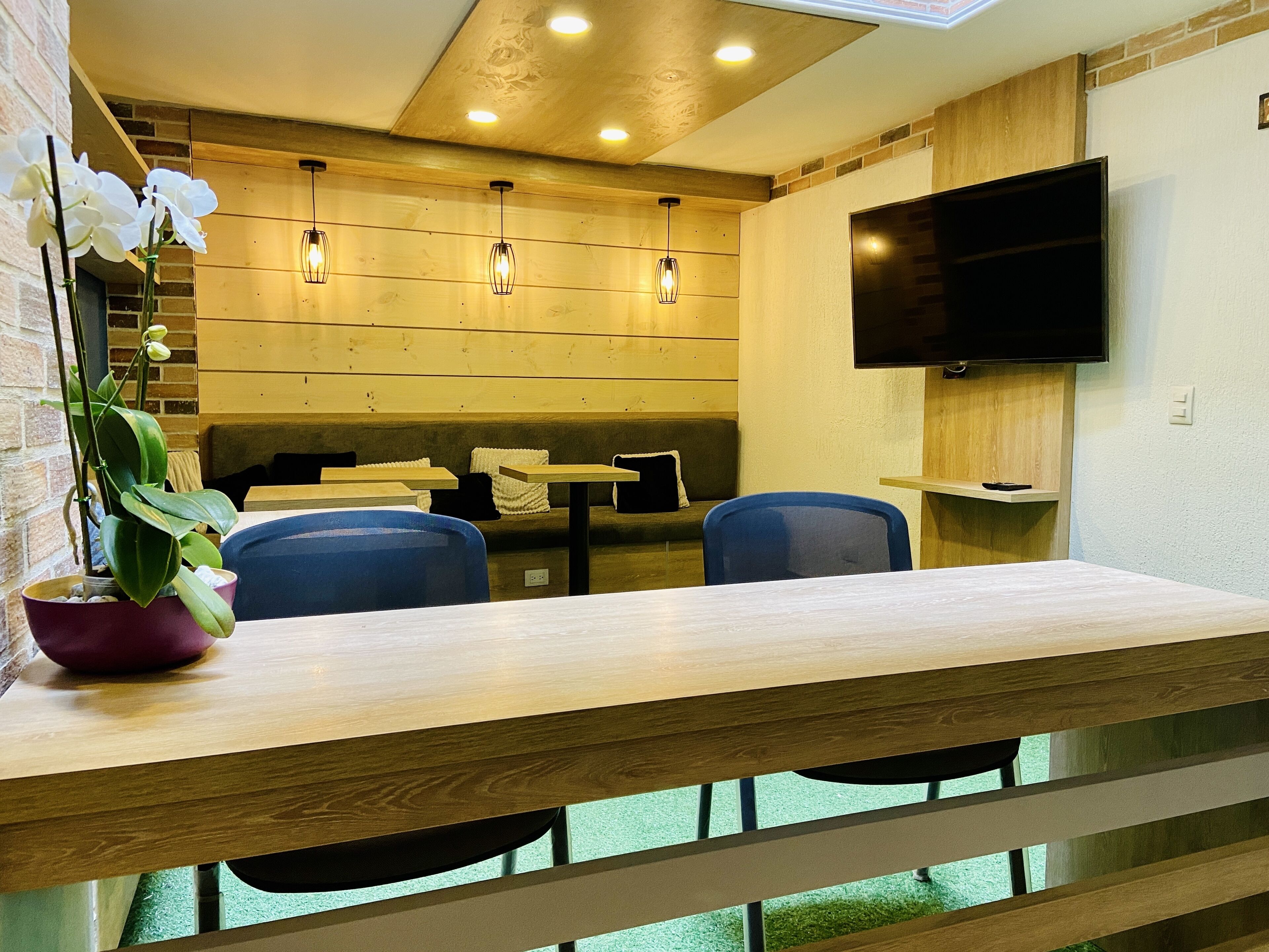A modern dining area with wooden tables and chairs, a potted plant, and a flat-screen TV mounted on the wall.