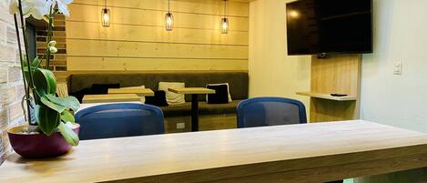 A modern dining area with wooden tables and chairs, a potted plant, and a flat-screen TV mounted on the wall.