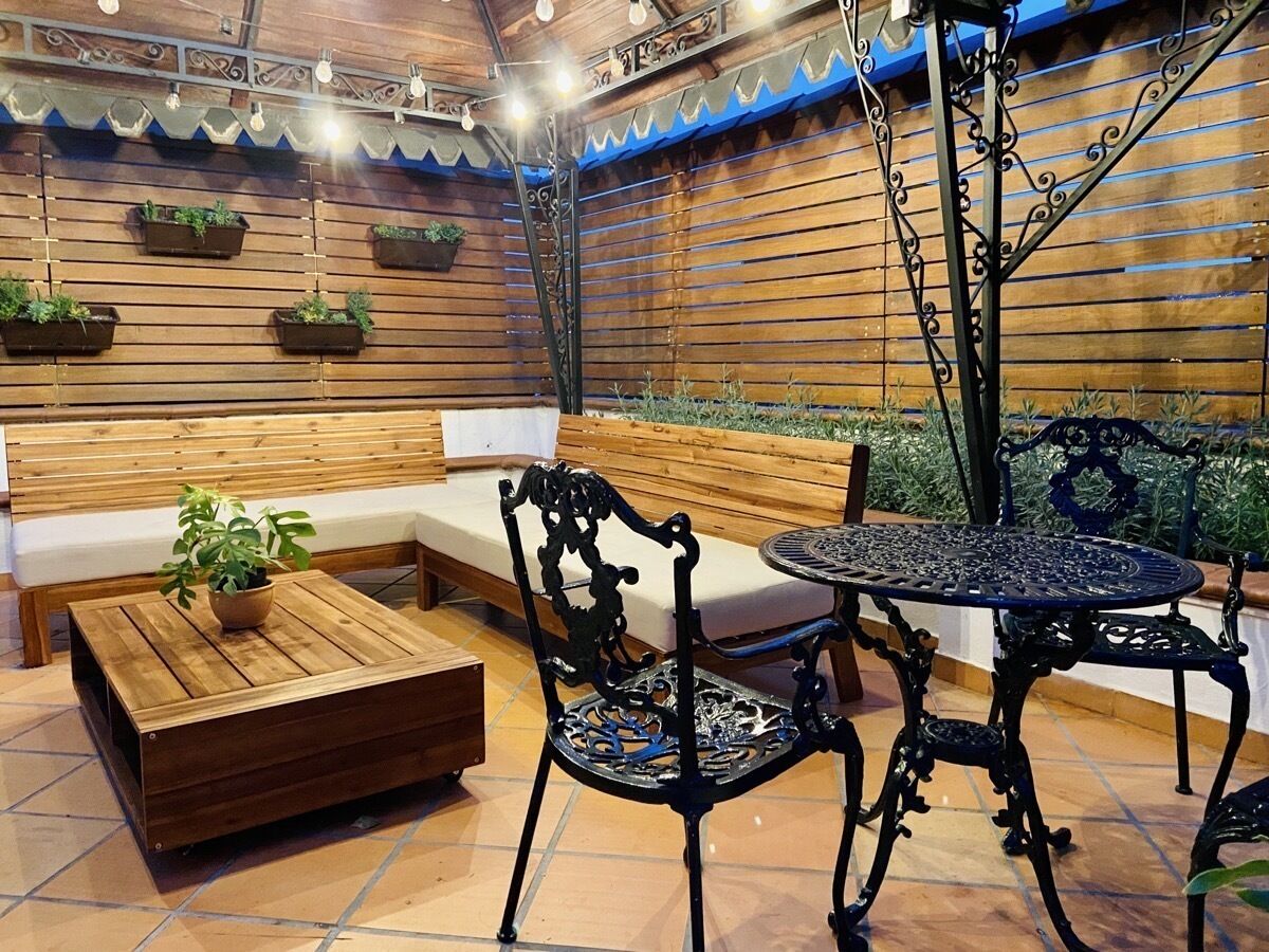 A wooden bench, a round metal table, and two chairs in a cozy outdoor seating area with hanging planters.