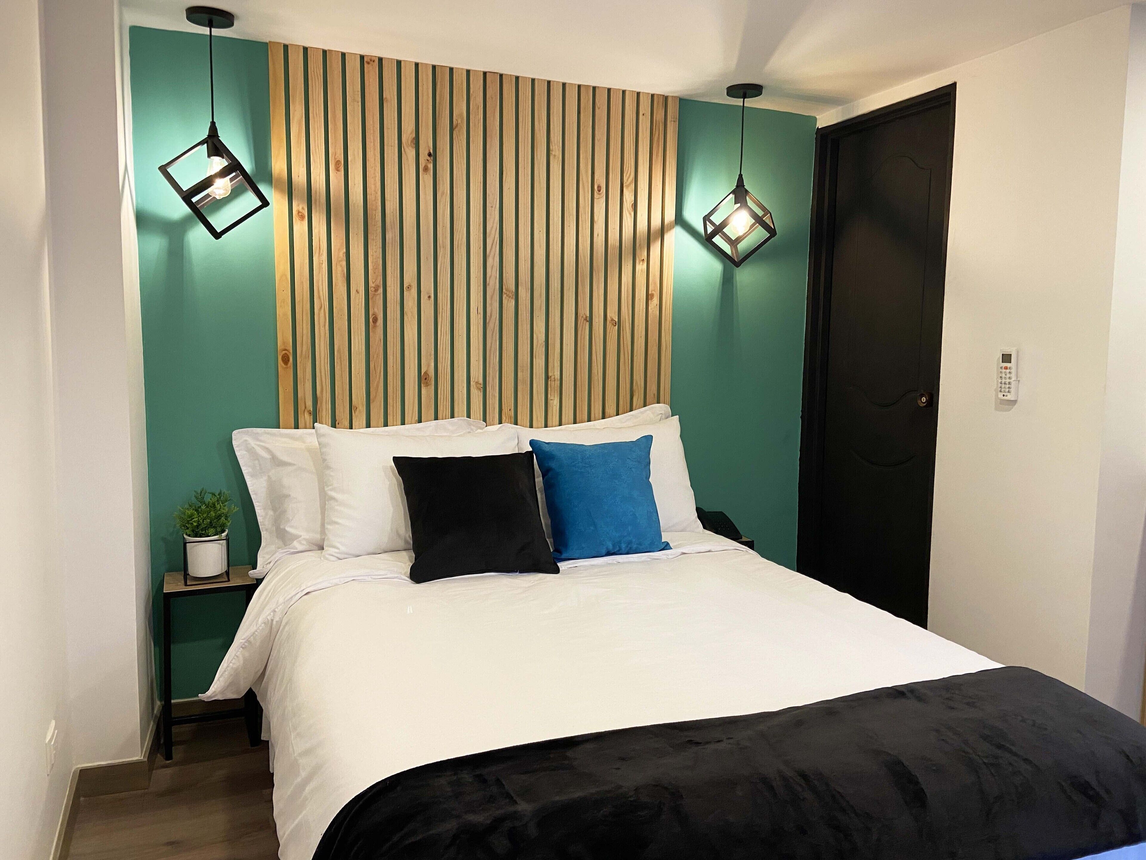 A bedroom with a bed, a wooden slat headboard, two pendant lights, a bedside table with a plant, and a door.