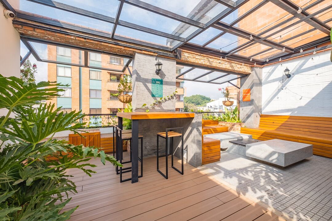 A rooftop patio with a wooden table and bar stools, surrounded by potted plants and a view of the cityscape.
