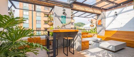 Una terraza en la azotea con una mesa de madera y taburetes tipo bar, rodeada de plantas en macetas y vistas panorámicas de la ciudad.