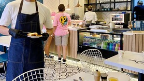 Interior de una cafetería moderna con un barista atendiendo, una barra con una máquina de café y un área de asientos con mesas y sillas.