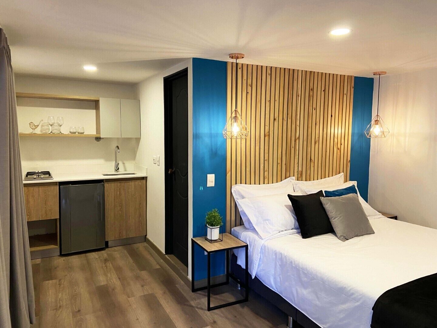 A compact hotel room with a bed, bedside table, and a kitchenette.