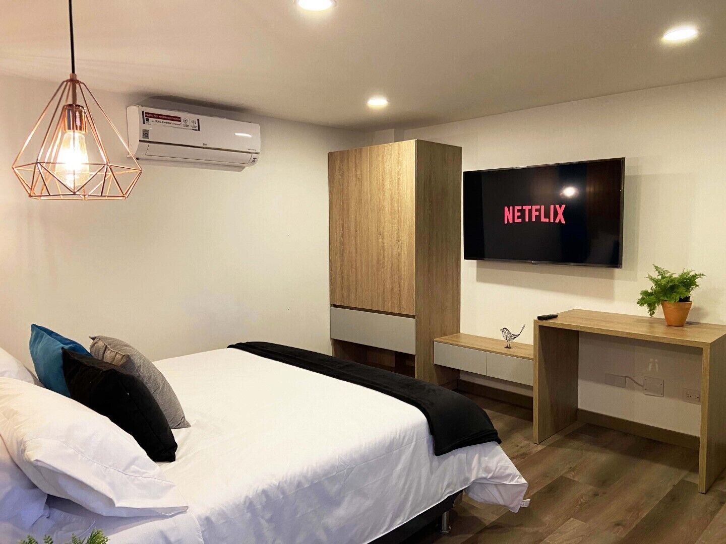 A modern hotel room with a bed, a desk, a TV displaying 'NETFLIX', and an air conditioning unit.