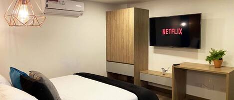 A modern hotel room with a bed, a desk, a TV displaying 'NETFLIX', and an air conditioning unit.