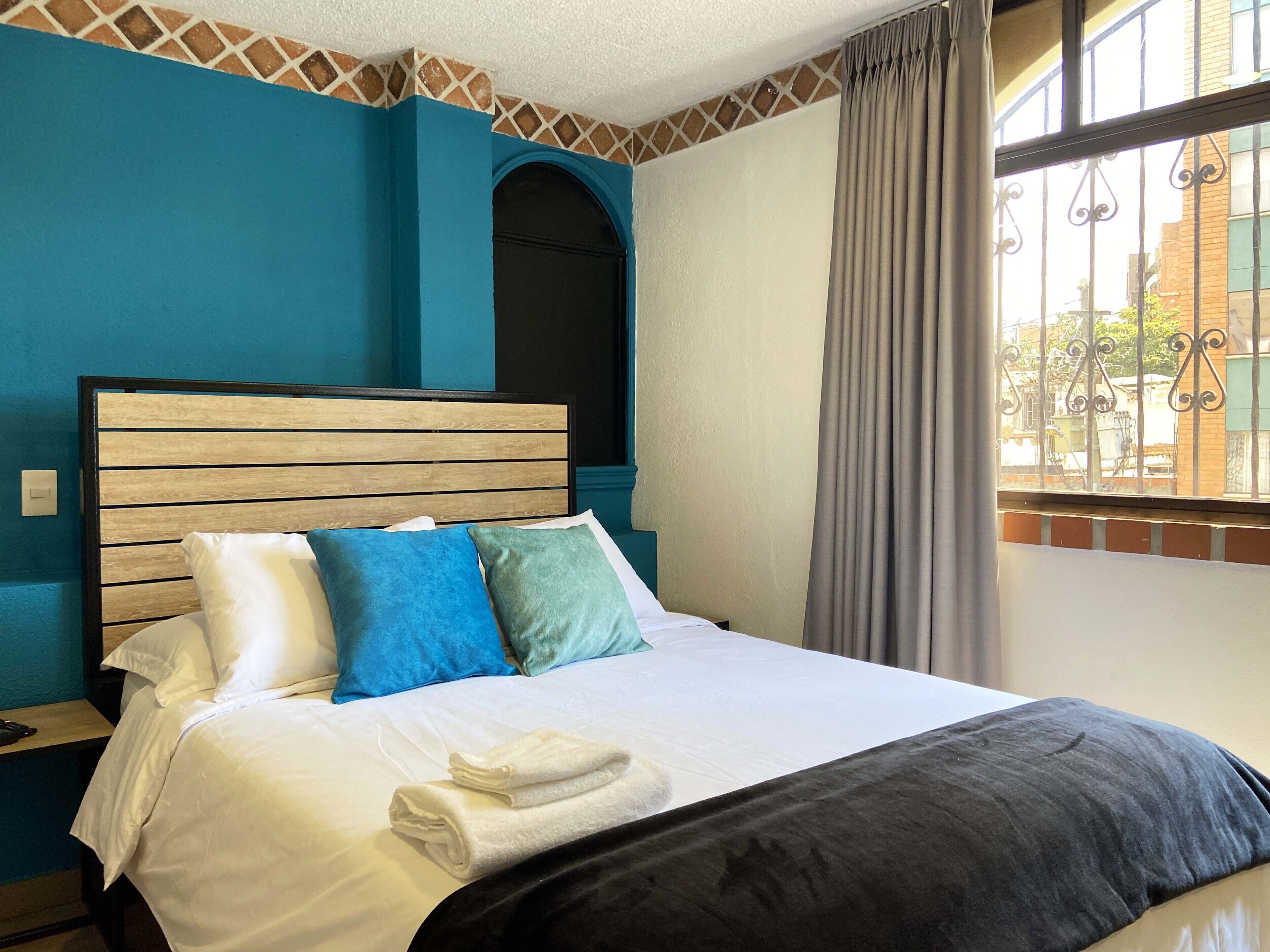 A hotel room with a wooden headboard, a bed with white and blue bedding, a window with a view of the outside, and a curtain.