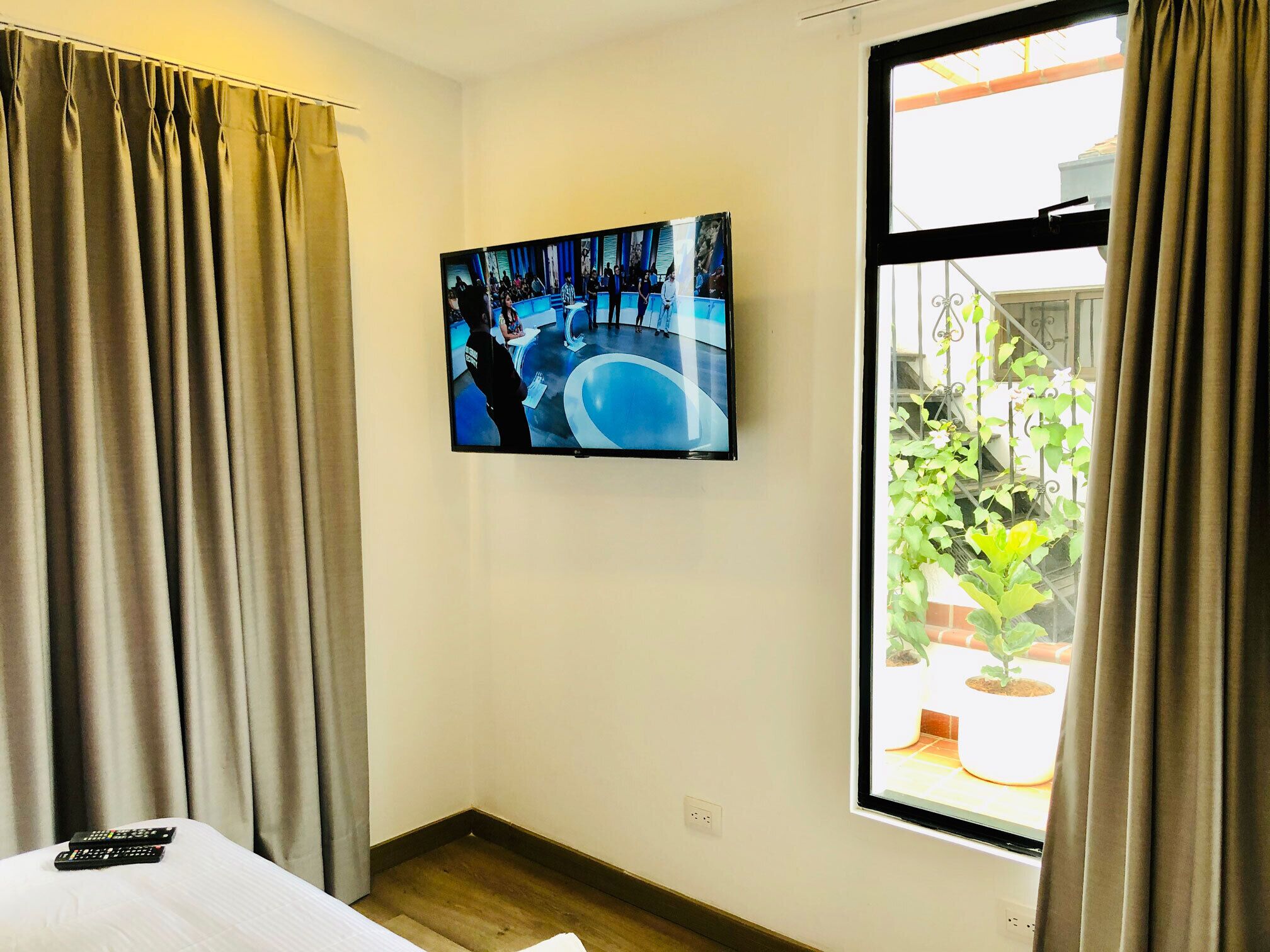 Een hotelkamer met een grote televisie aan de muur, een raam met uitzicht op een balkon en een bed met een witte dekbedovertrek.