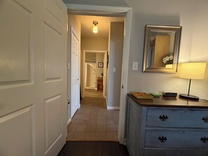 Un couloir avec un plancher en bois, une porte blanche sur la gauche, une commode avec des tiroirs, un miroir et une lampe sur la droite.