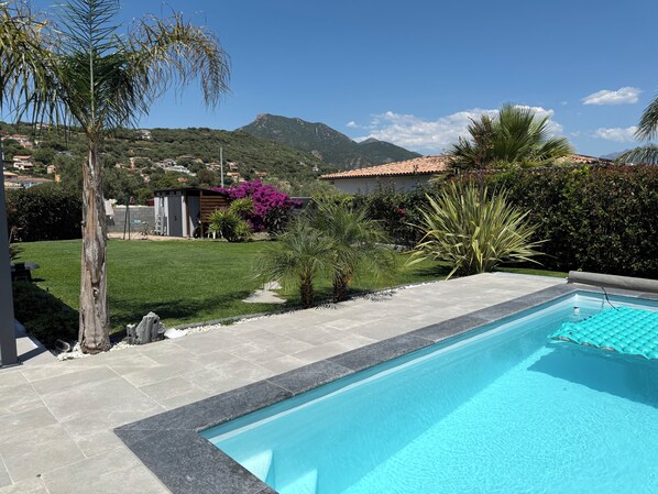 Una piscina con borde de azulejos, rodeada de césped, palmeras y una montaña al fondo.