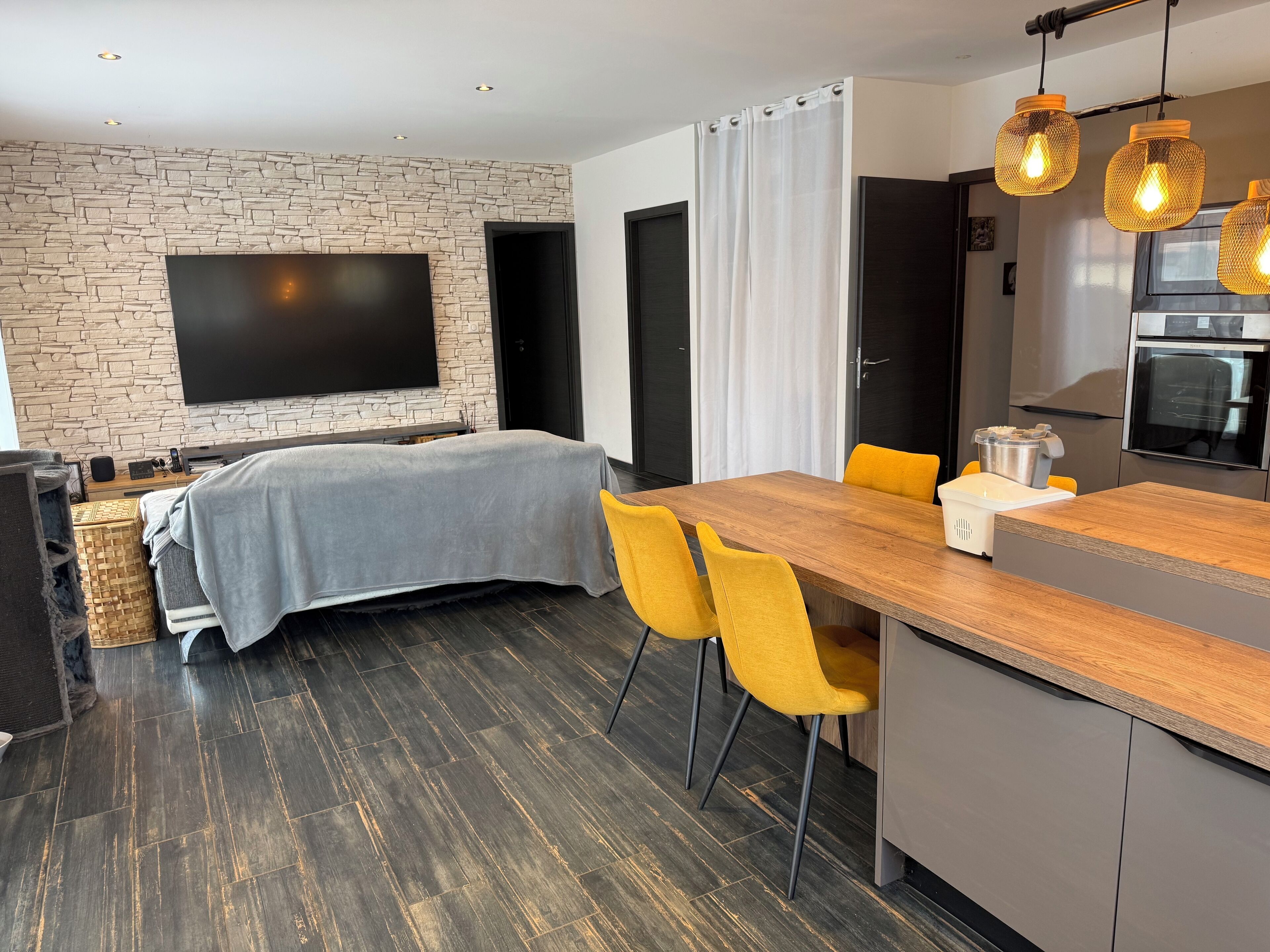 Een moderne keuken met een houten aanrecht, gele stoelen, een ingebouwde oven en een grote flatscreen televisie die aan een stenen muur is bevestigd.