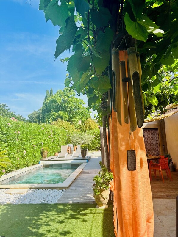 A pool area with a wooden deck, a pergola with hanging vines, and a view of a lush garden.