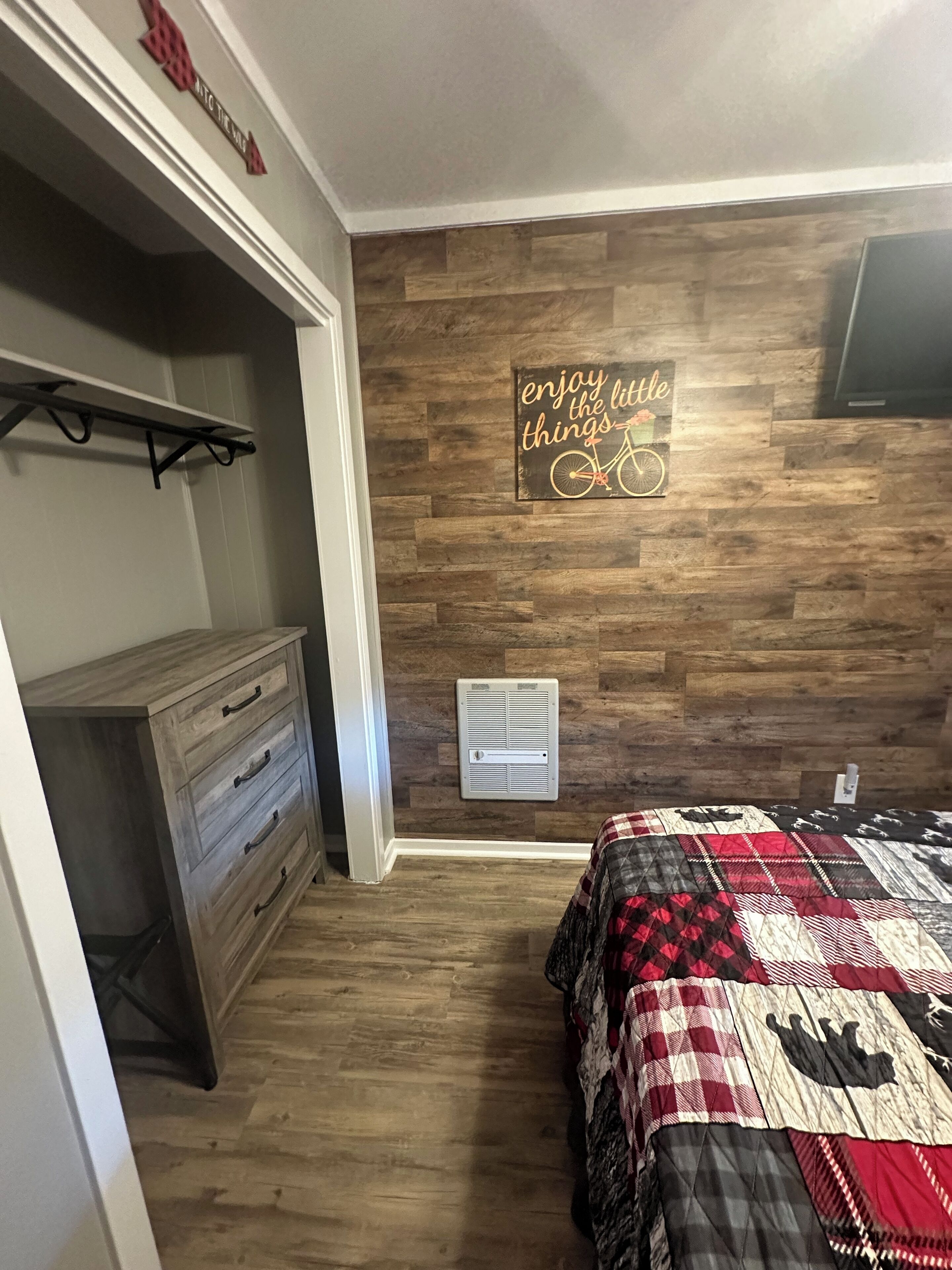 A bedroom with a wooden wall, a bed with a quilt, a dresser, and a wall-mounted air vent.