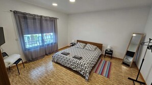 Une chambre à coucher comprenant un lit, une chaise, une table de chevet, une lampe, un miroir et une fenêtre avec des rideaux.
