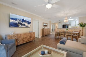 A spacious living room with a large flat-screen TV, a dining area with a wooden table and chairs, and a kitchen in the background. - Ocean Reef 104 - intimate condo with pool (St Augustine Beach)