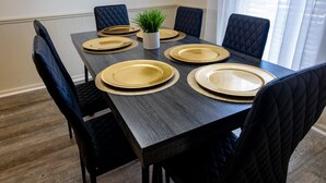 Une table à manger dressée avec de l’argenterie dorée et des chaises noires, une petite plante dans un pot blanc et une fenêtre ornée de rideaux légers.