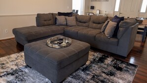 Un salon moderne avec un canapé d’angle, un pouf assorti et un tapis à motifs.