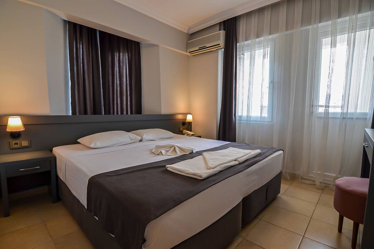A hotel room with a large bed, two bedside lamps, an air conditioning unit, a desk, and a chair.