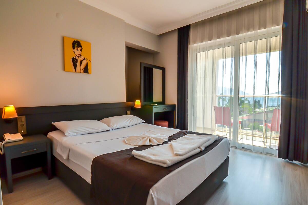 A hotel room with a large bed, a desk, a chair, a lamp, a telephone, a mirror, and a window with curtains.