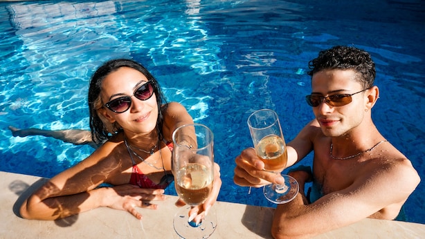 Zwei Personen in einem Schwimmbecken, die Gläser mit einer klaren Flüssigkeit, vermutlich einem Getränk, halten.